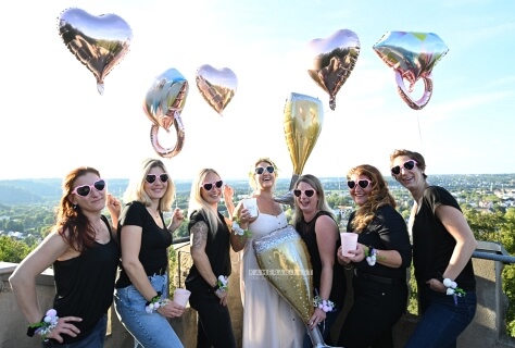Foto kurz vor Sonnenuntergang: Die Braut inmitten ihrer gut gelaunten Freundinnen im schönen Abendlicht. Blumenarmbänder, Herzchenbrillen und Heliumballons schmücken die Freundinnen.